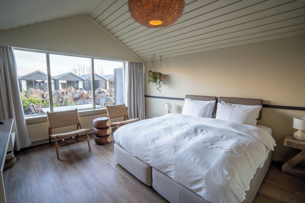 Une chambre avec un grand lit, une fenêtre donnant sur des immeubles, un luminaire suspendu en osier et une chaise près de la fenêtre.