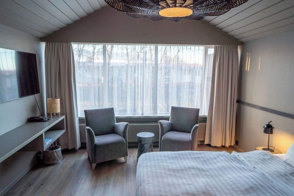 Een hotelkamer met een bed, twee fauteuils, een klein rond tafeltje, een televisie op een plank en een groot raam met gordijnen.