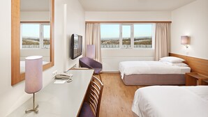Une chambre d’hôtel avec un grand miroir, un bureau avec une lampe, deux lits, une télévision et une fenêtre donnant sur la ville.