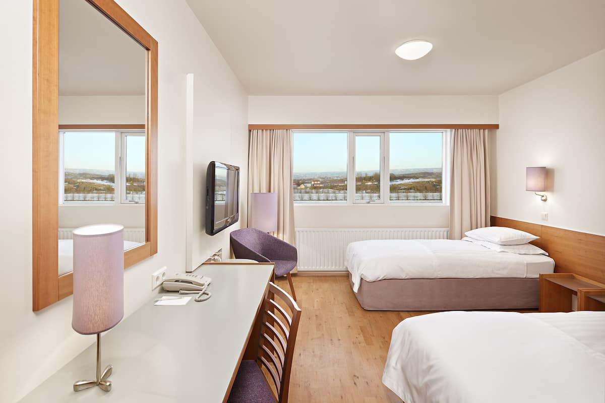 Ein Hotelzimmer mit einem großen Spiegel, einem Schreibtisch mit Lampe, zwei Betten, einem Fernseher und einem Fenster mit Stadtblick.