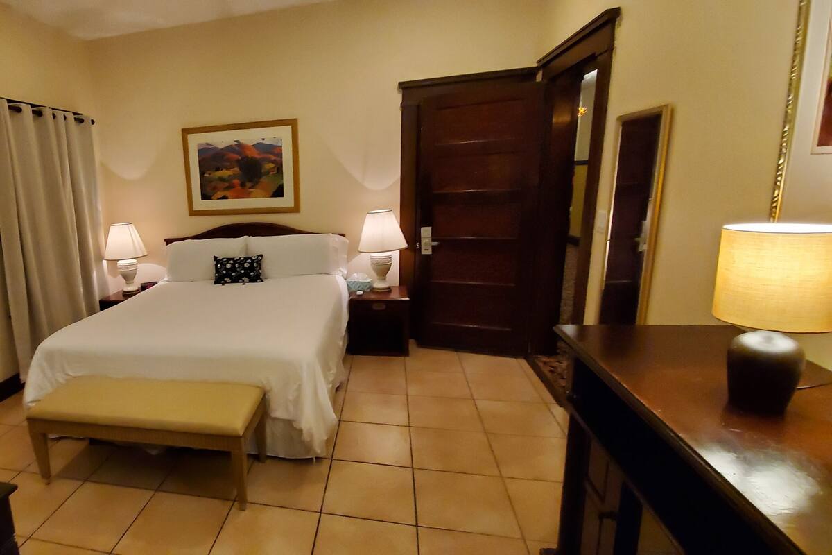 A hotel room with a bed, two bedside tables with lamps, a bench, a door, and a painting on the wall.