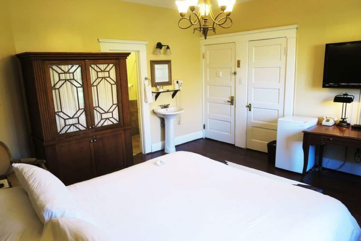 A hotel room with a large bed, a wooden wardrobe, a white sink, a television, and a desk with a lamp.