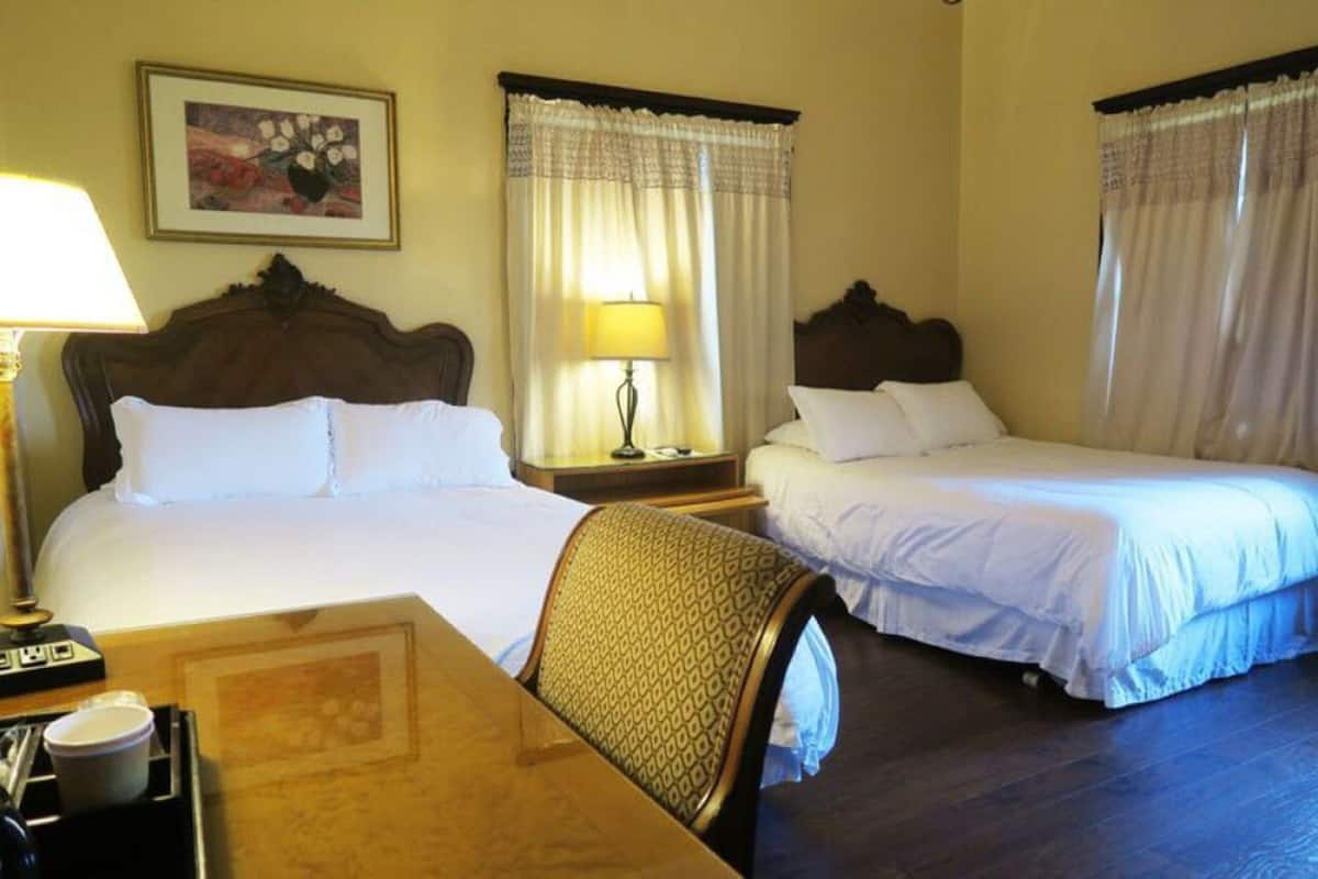 A hotel room with two single beds, a wooden table with a cup and a tray, a chair, and a lamp.