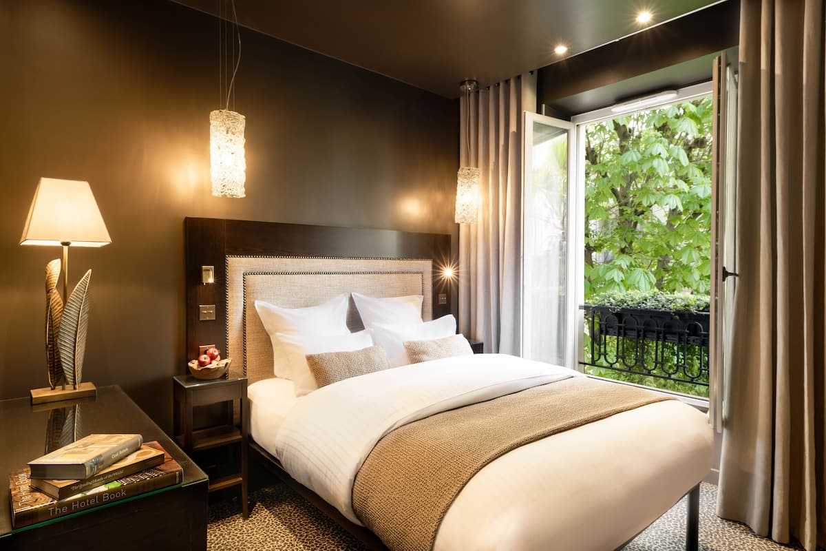 Quarto de hotel moderno com uma cama grande, mesa de cabeceira com abajur e vista para o verde pela janela.