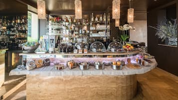 A bar area with a marble counter, various bottles, glasses, and a selection of fruits and pastries.