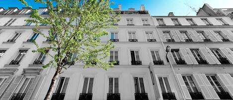 A multi-story building with white facades, multiple windows with shutters, and a tree in the foreground.