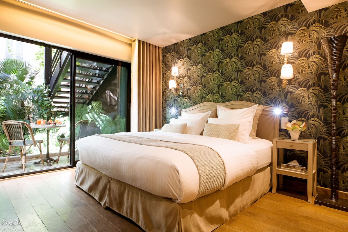 Quarto com cama grande, mesa de cabeceira e vista para uma varanda com plantas e escadas.