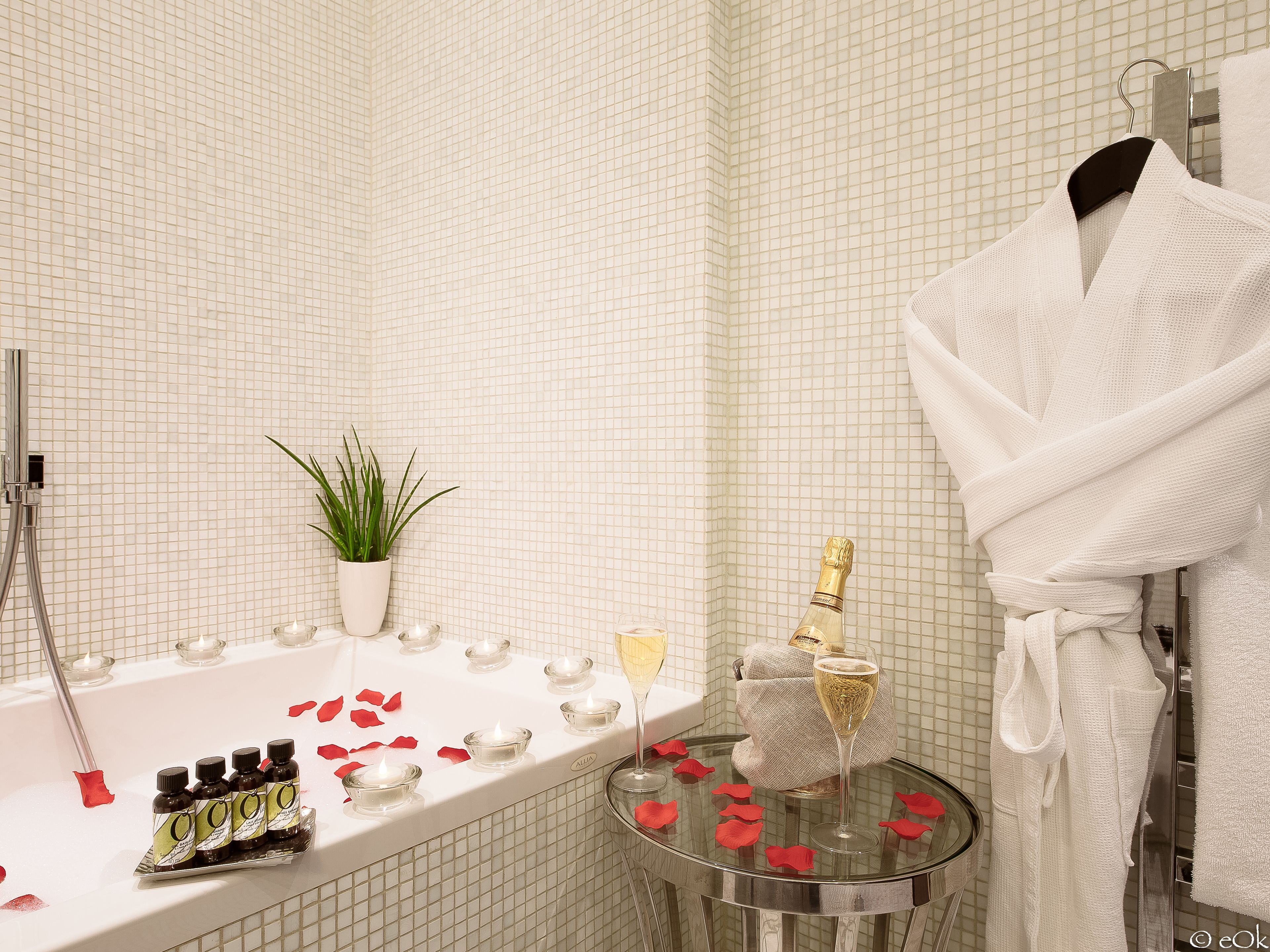 A bathroom with a bathtub, a glass table with candles, a bottle of champagne, and a robe hanging on a rack.