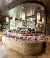 Uma área de bar com balcão de mármore, diversos alimentos e bebidas, e uma estante bem abastecida com garrafas e copos.