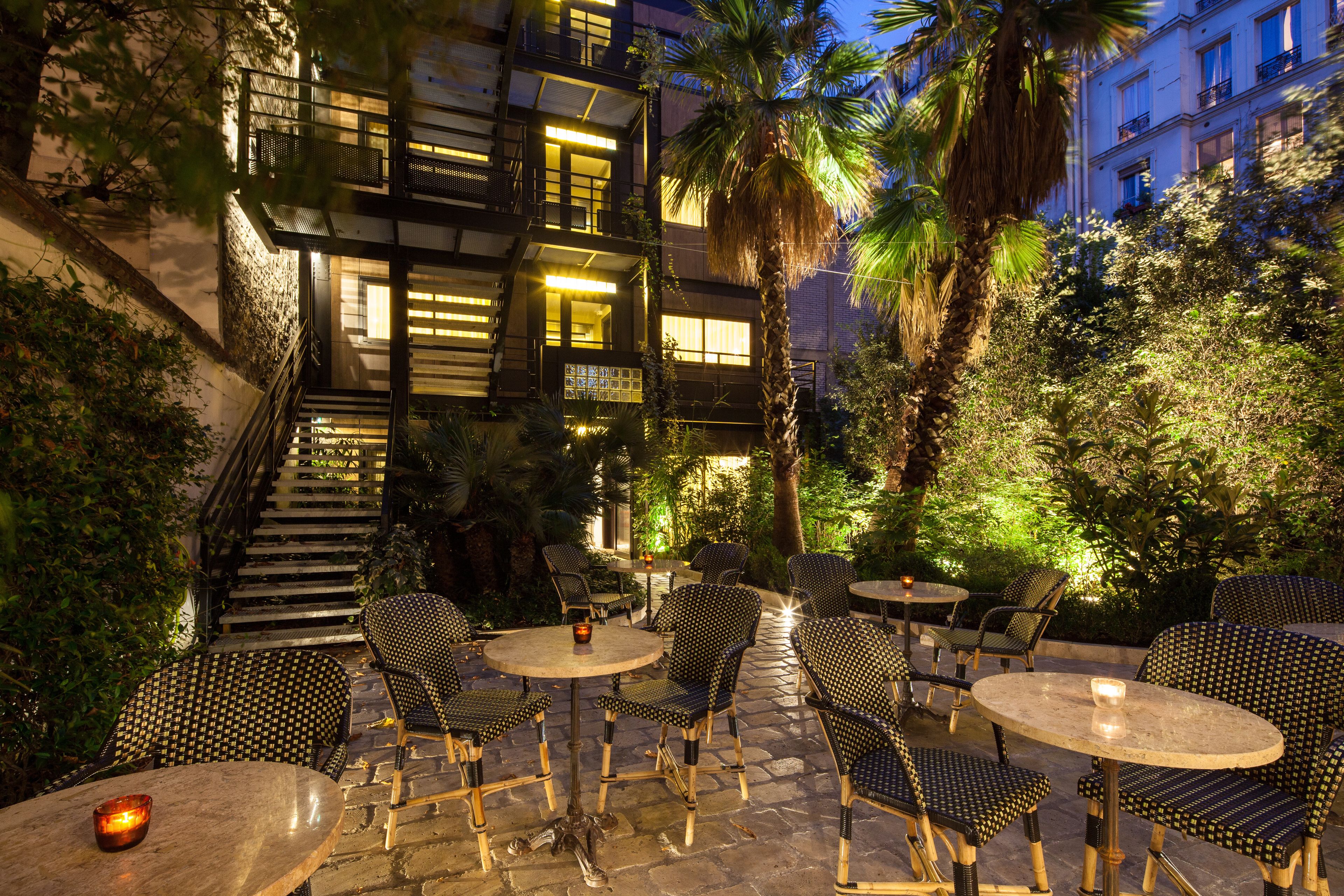 An outdoor patio with wicker furniture and a stone floor, surrounded by lush greenery and a staircase leading to an upper level.