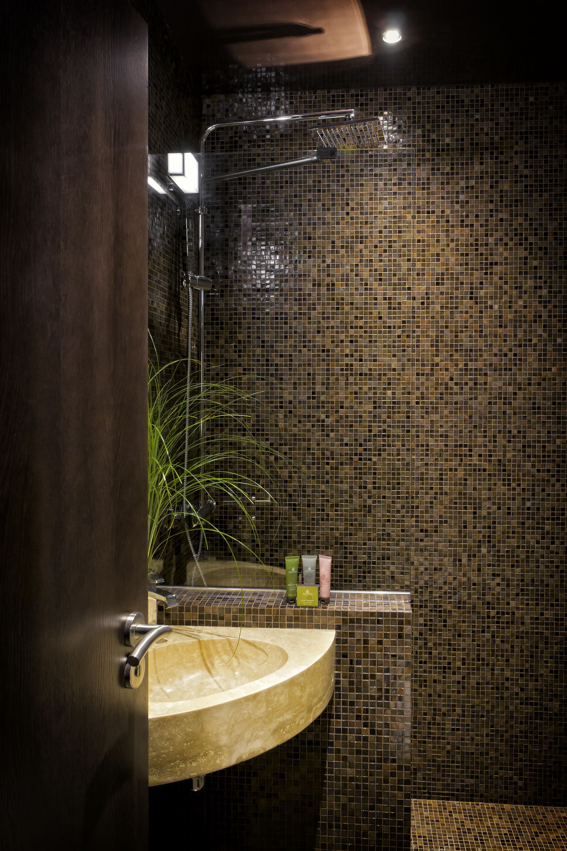 Un baño con pared de mosaicos, puerta de madera, lavamanos y zona de ducha.