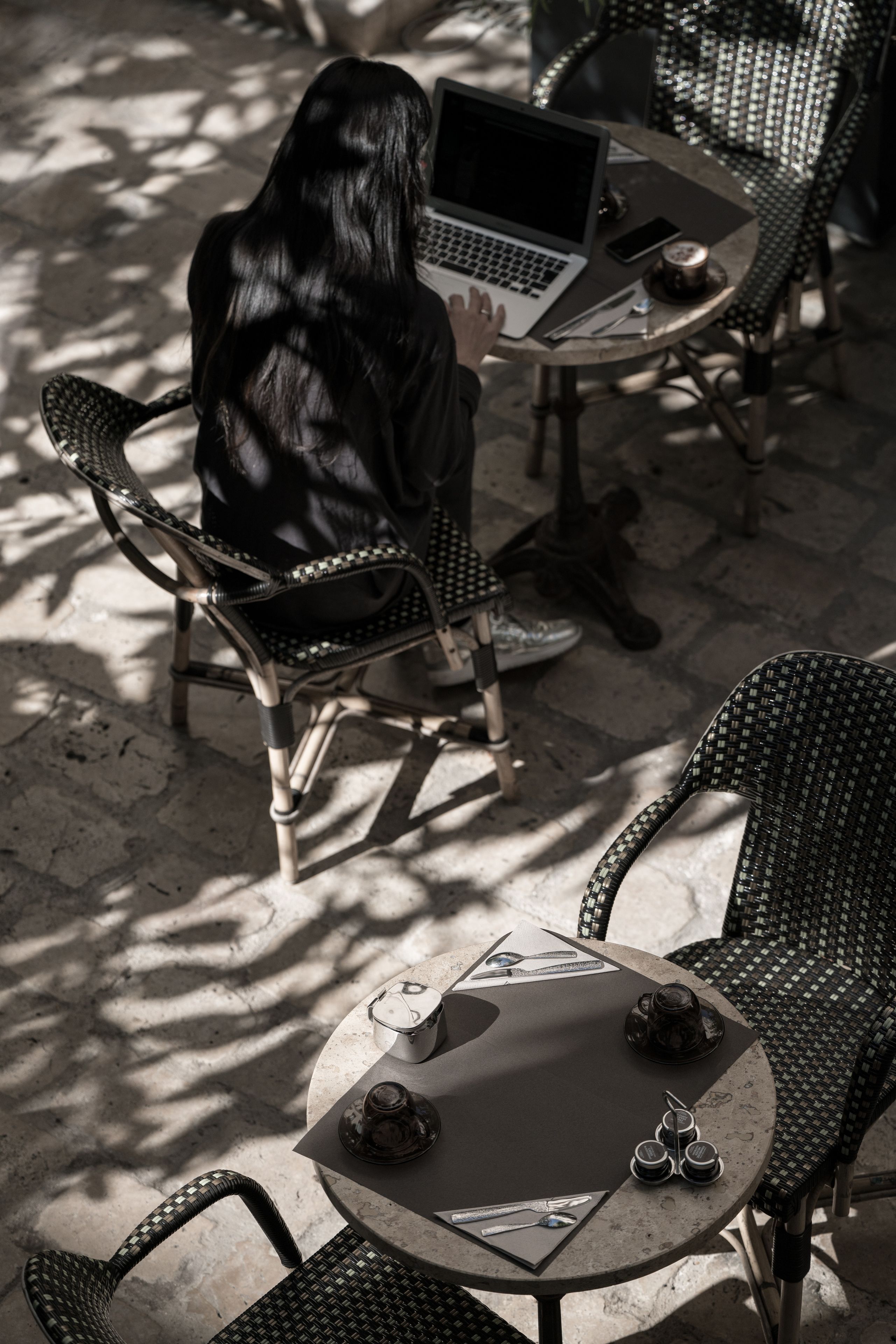 Une personne est assise à une table à l’extérieur, devant un ordinateur portable, avec une tasse et un téléphone intelligent posés sur la table.