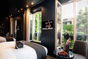 Quarto de hotel com cama, mesa de cabeceira com uma taça de morangos, uma taça de vinho e um aviso indicando o local como Moulin Rouge.