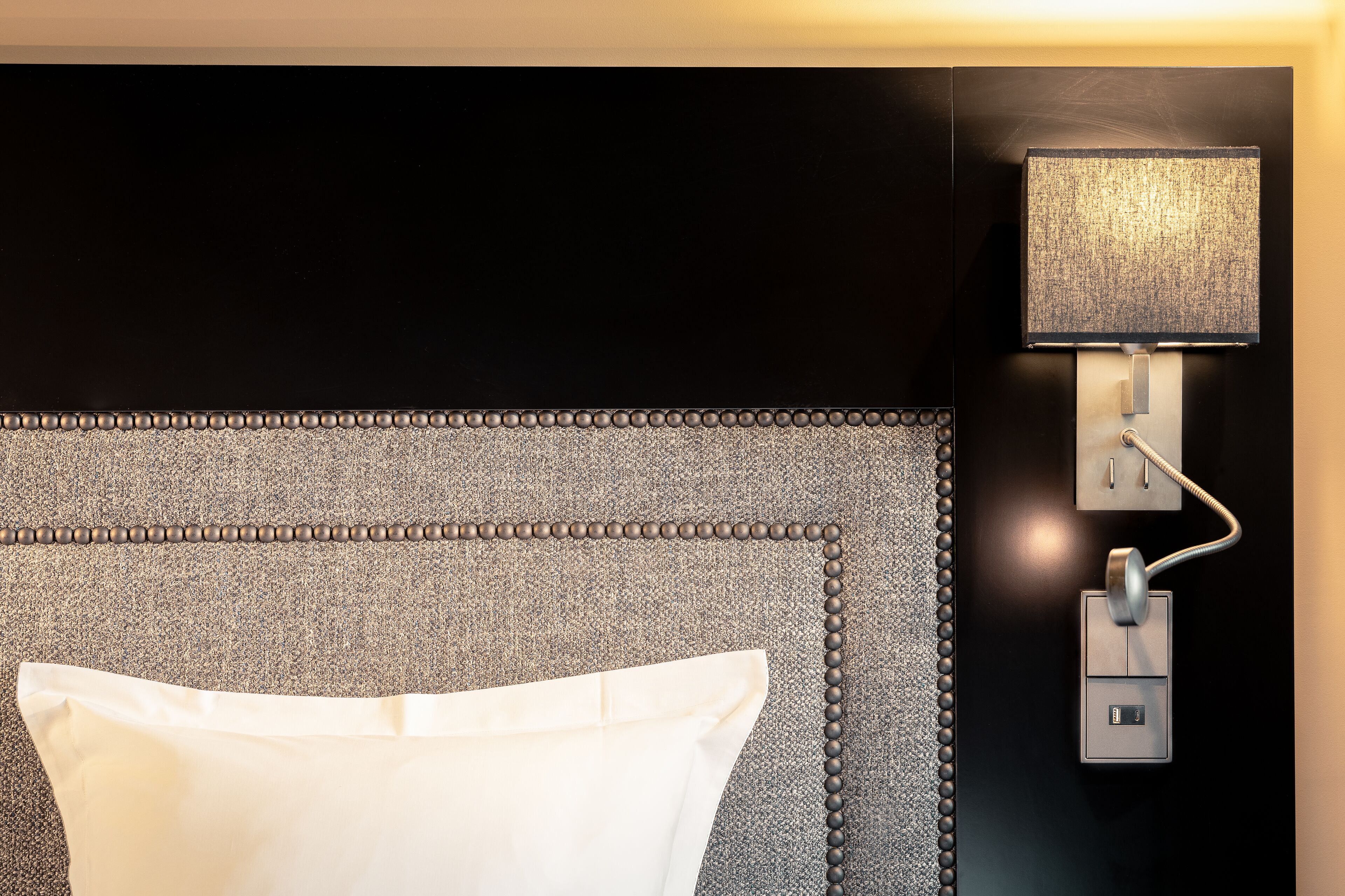 A modern hotel room with a textured headboard, a bedside lamp, and a control panel.