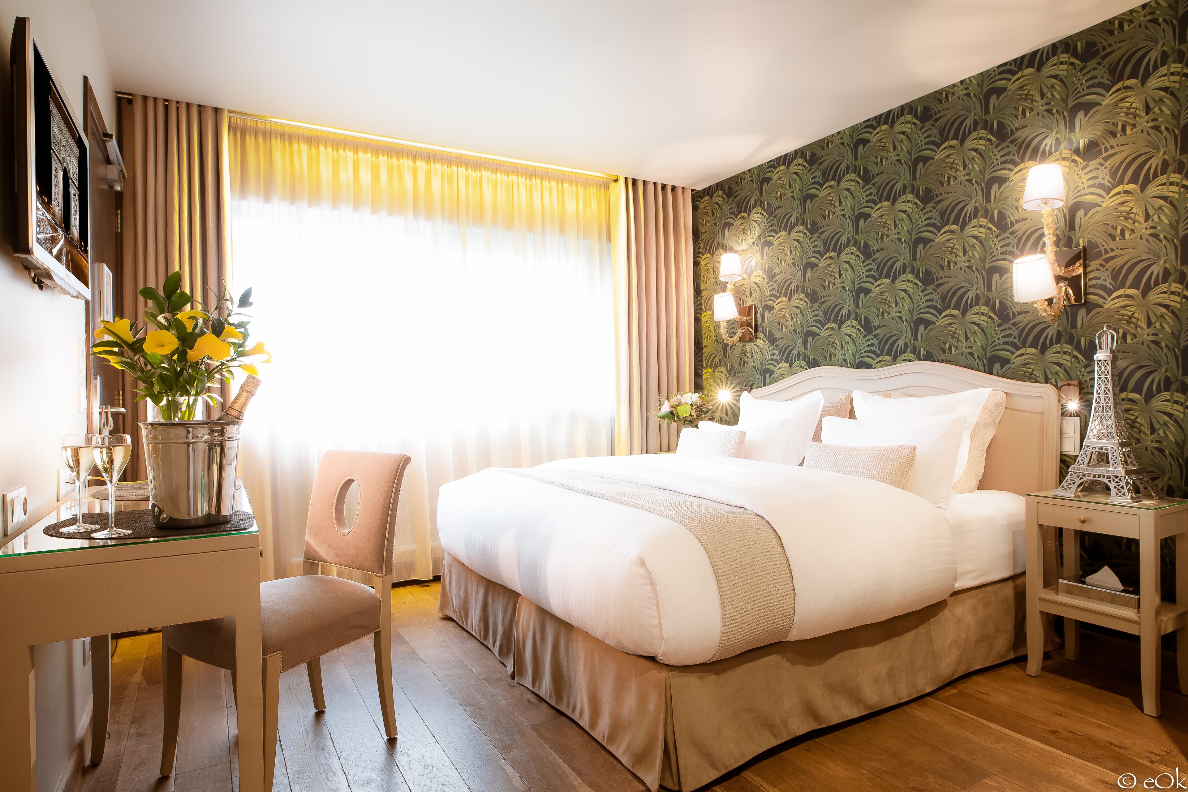 A hotel room with a large bed, a desk with a chair, a vase with flowers, and a nightstand with a lamp.
