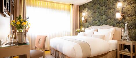 A hotel room with a large bed, a desk with a chair, a vase with flowers, and a nightstand with a lamp.