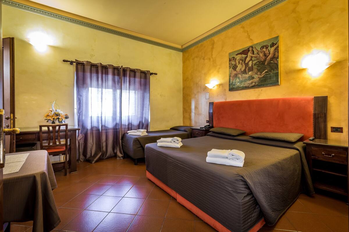 A hotel room with a large bed, a desk, and a chair. There is a painting on the wall and a window with curtains.