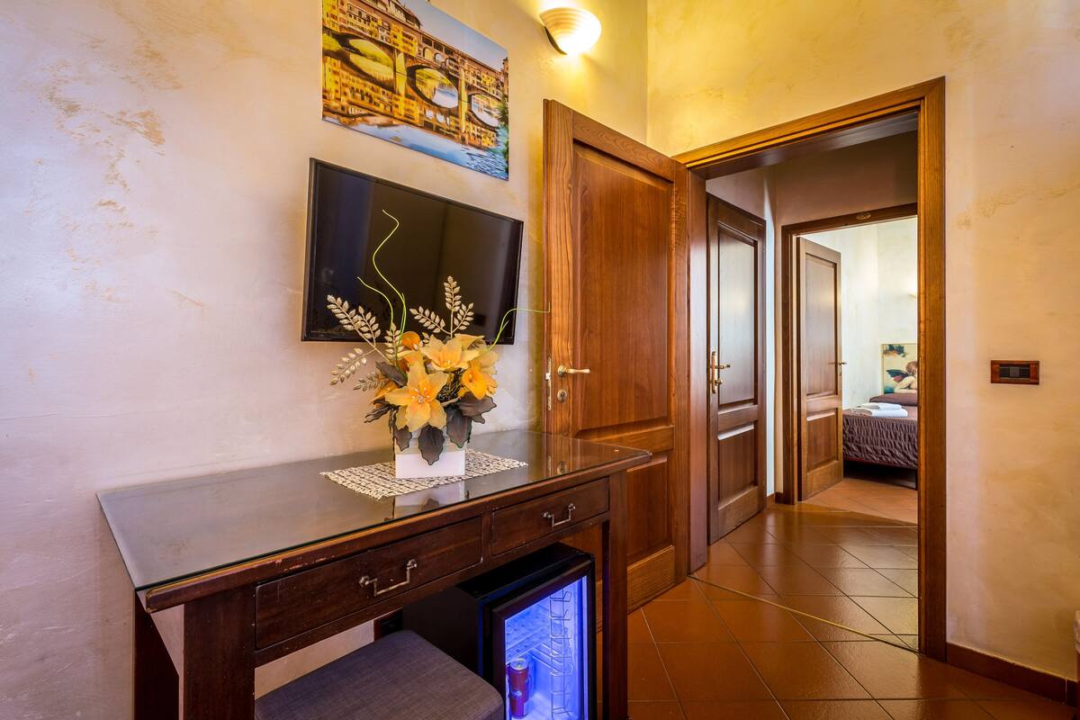 A hallway with a wooden door, a small refrigerator, a glass table with a vase of flowers, and a wall-mounted picture.