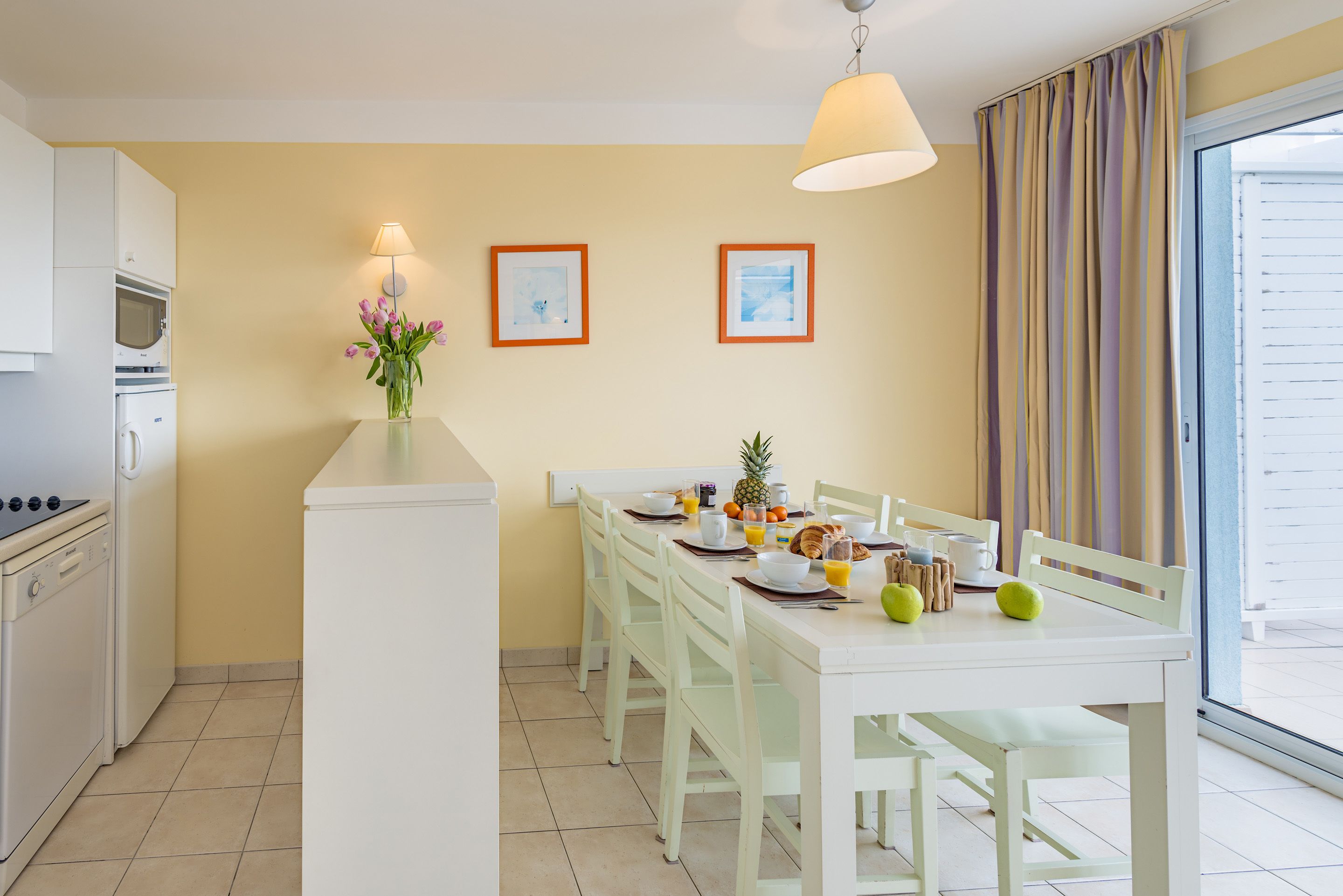 Une cuisine avec des meubles blancs, une table à manger avec des chaises, un vase rempli de fleurs et deux tableaux encadrés accrochés au mur.