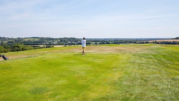 Una persona parada en un campo cubierto de hierba, con un palo de golf, observando un paisaje con campos y edificios a la distancia.