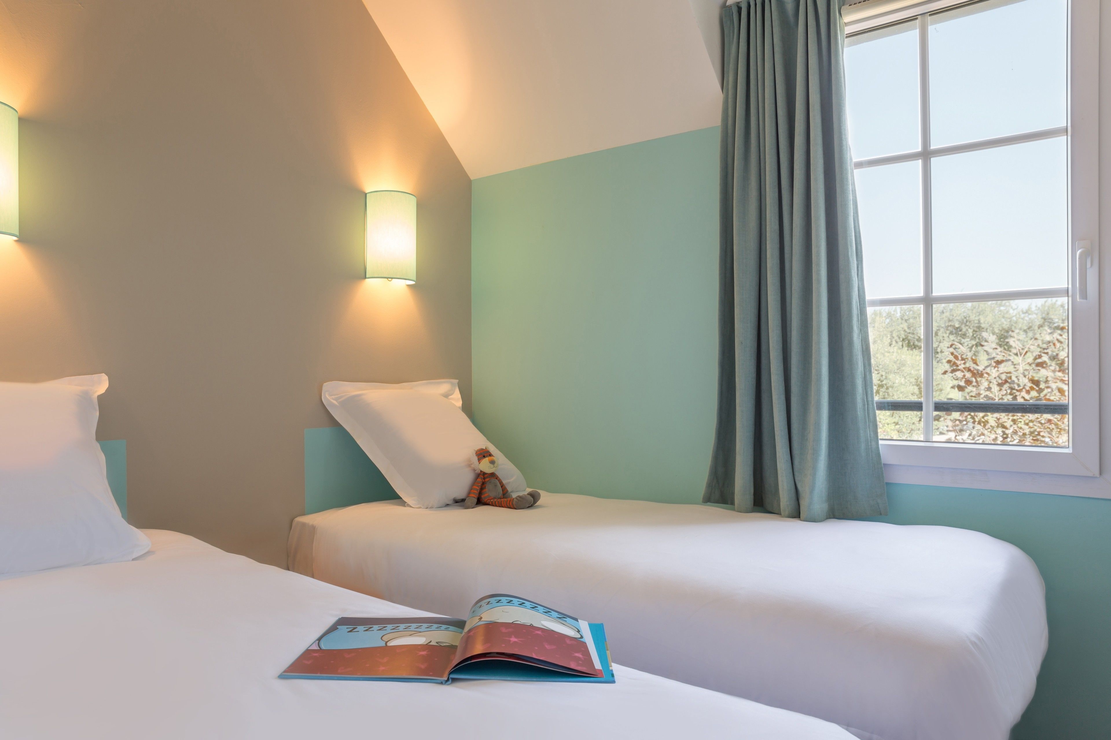 A hotel room with two beds, a window with curtains, and wall-mounted lights.