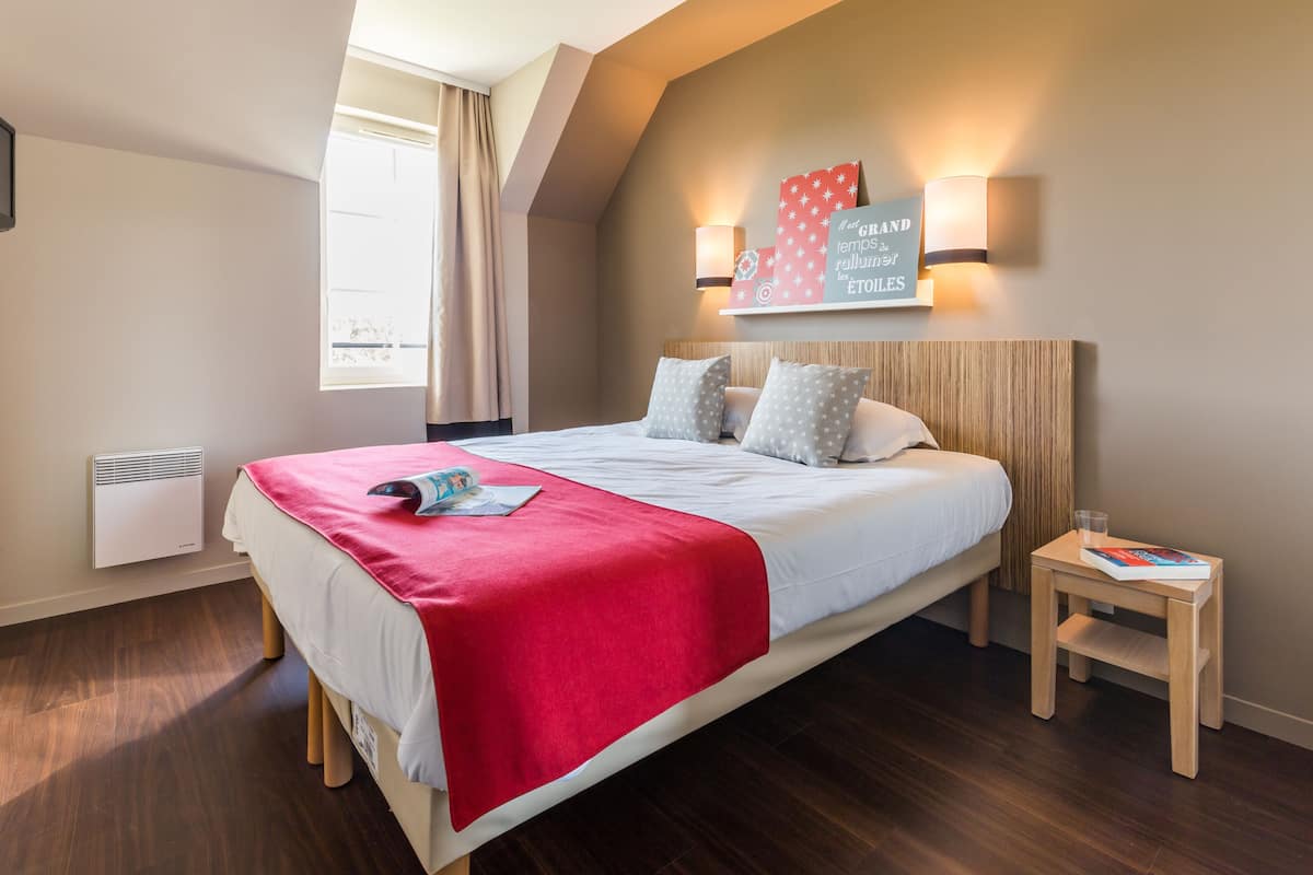 A bedroom with a bed, a nightstand, a wall-mounted light, a window with curtains, and a wall decoration.