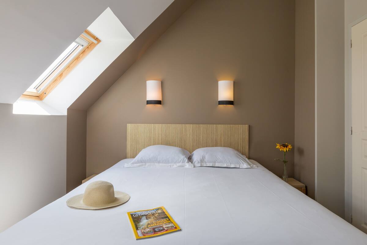 A bedroom with a sloped ceiling, skylight, two bedside lamps, a bed with white linens, a hat, and a magazine on the bed.