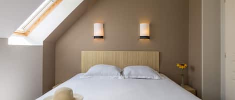 Ein Schlafzimmer mit schrägem Dach, Oberlicht, zwei Nachttischlampen, einem Bett mit weißen Leinen, einem Hut und einer Zeitschrift auf dem Bett.