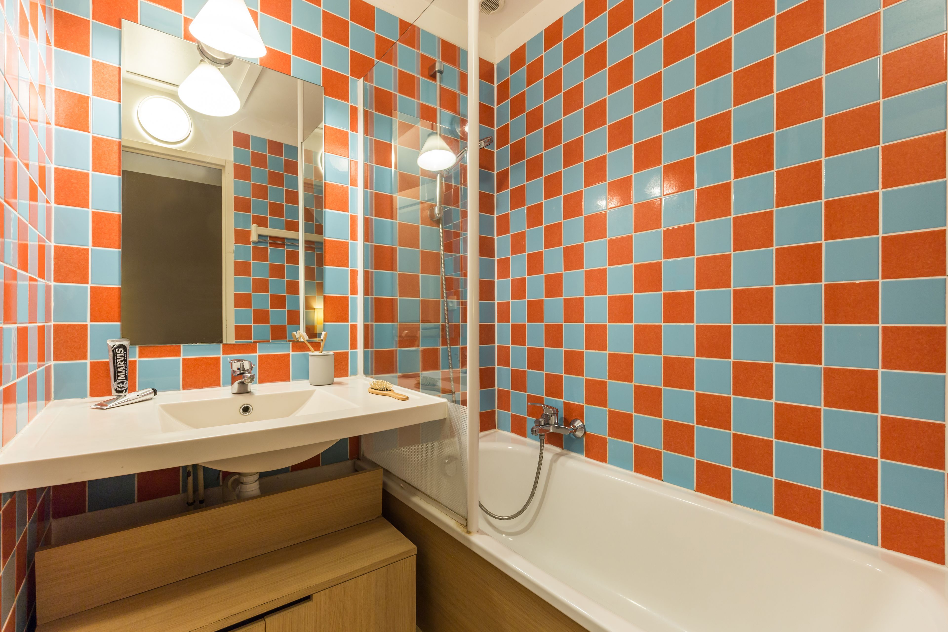 A bathroom with a white sink, mirror, and bathtub. The walls are tiled with a checkered pattern of orange and blue tiles.