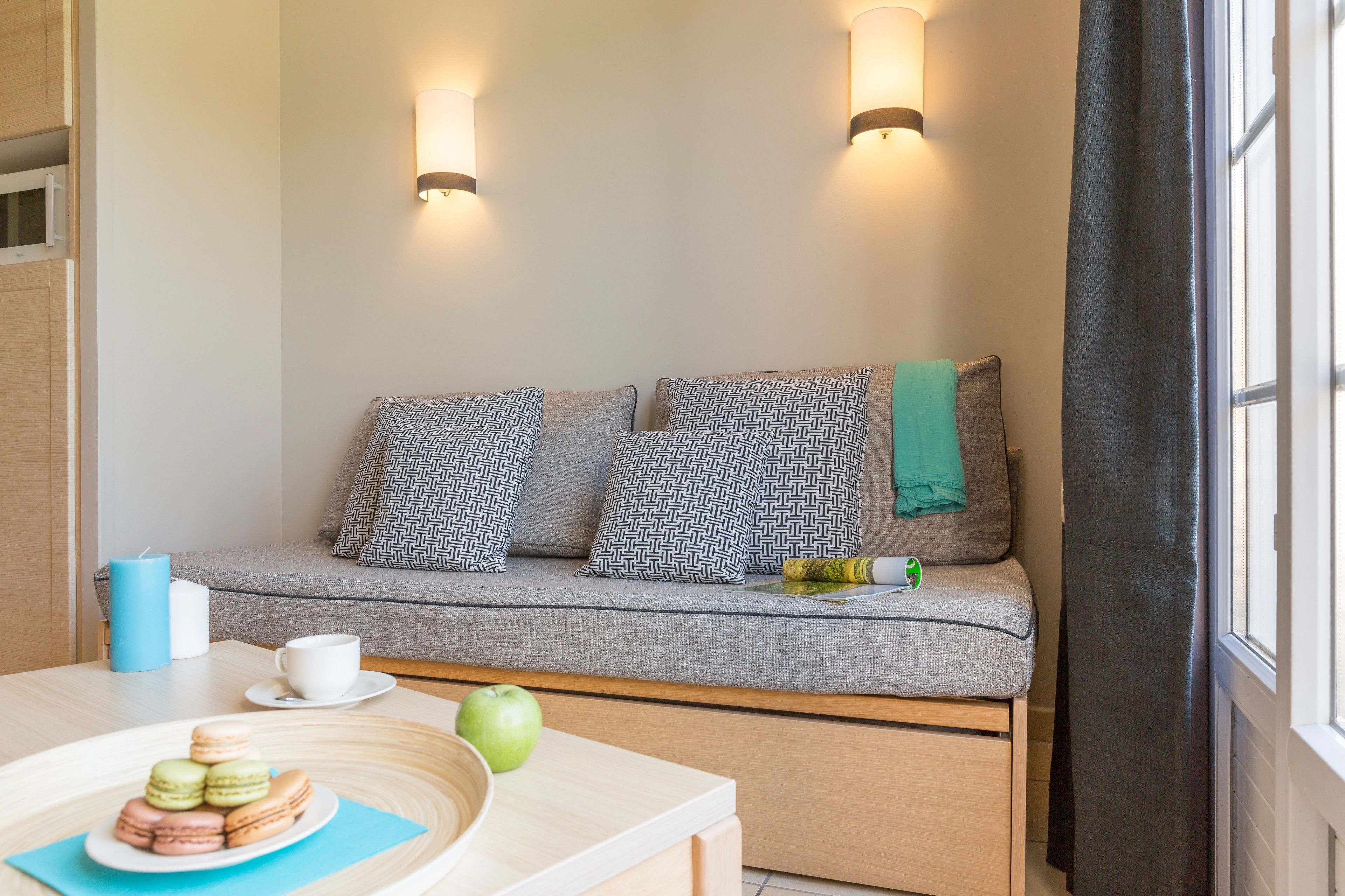 A cozy room with a sofa, pillows, a table with a plate of macarons and an apple, and a window with curtains.