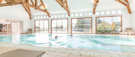 En indendørs swimmingpool med træbjælker, store vinduer og liggestole.