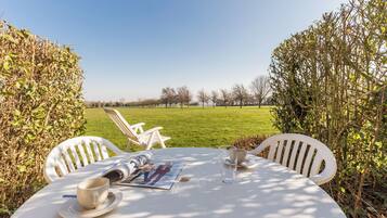 Ein runder Tisch mit einer Zeitschrift, einer Tasse und einem Glas, draußen in der Sonne, mit weißen Stühlen und Blick auf ein grünes Feld.