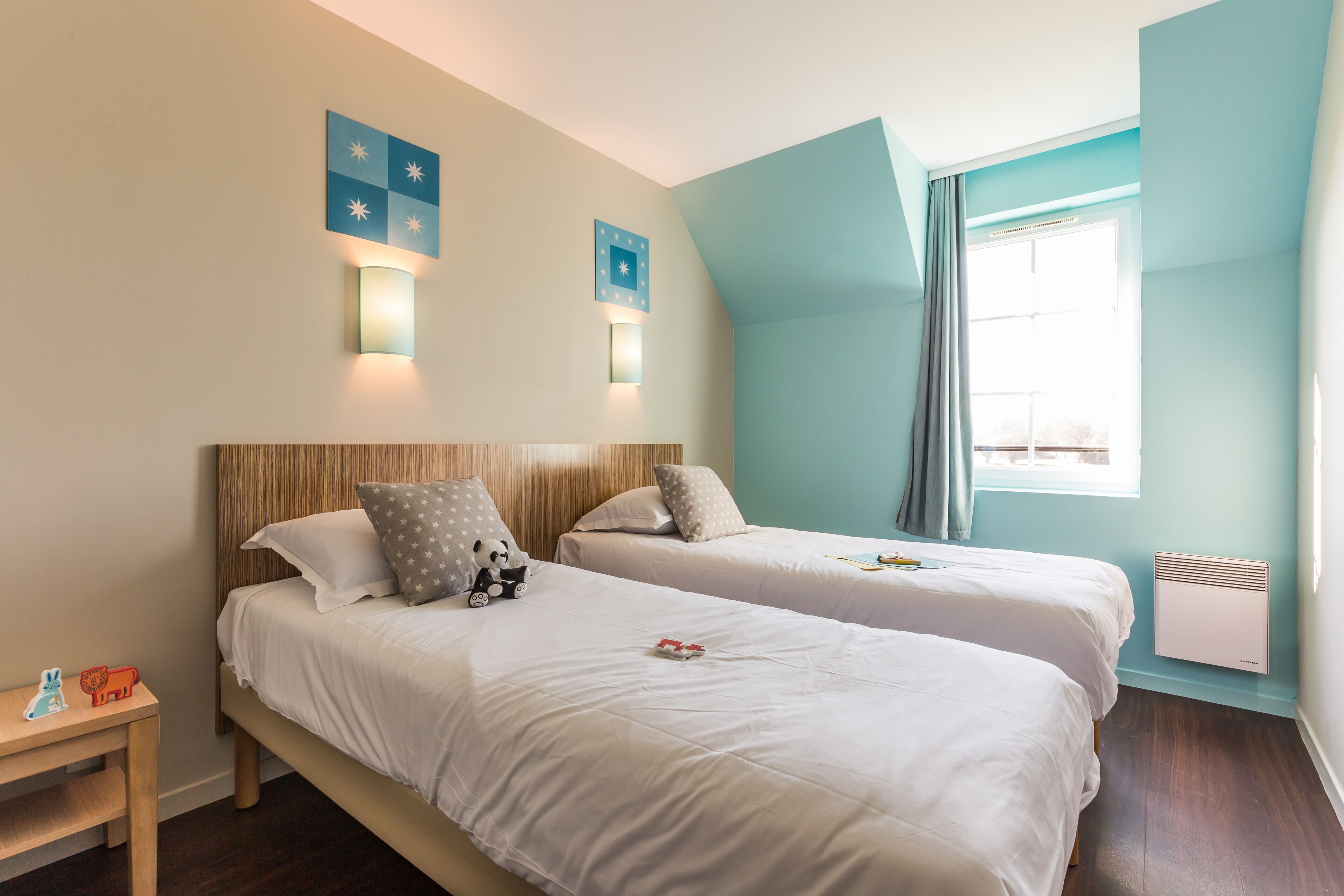 A hotel room with two beds, a nightstand, a window with curtains, and wall-mounted lights.