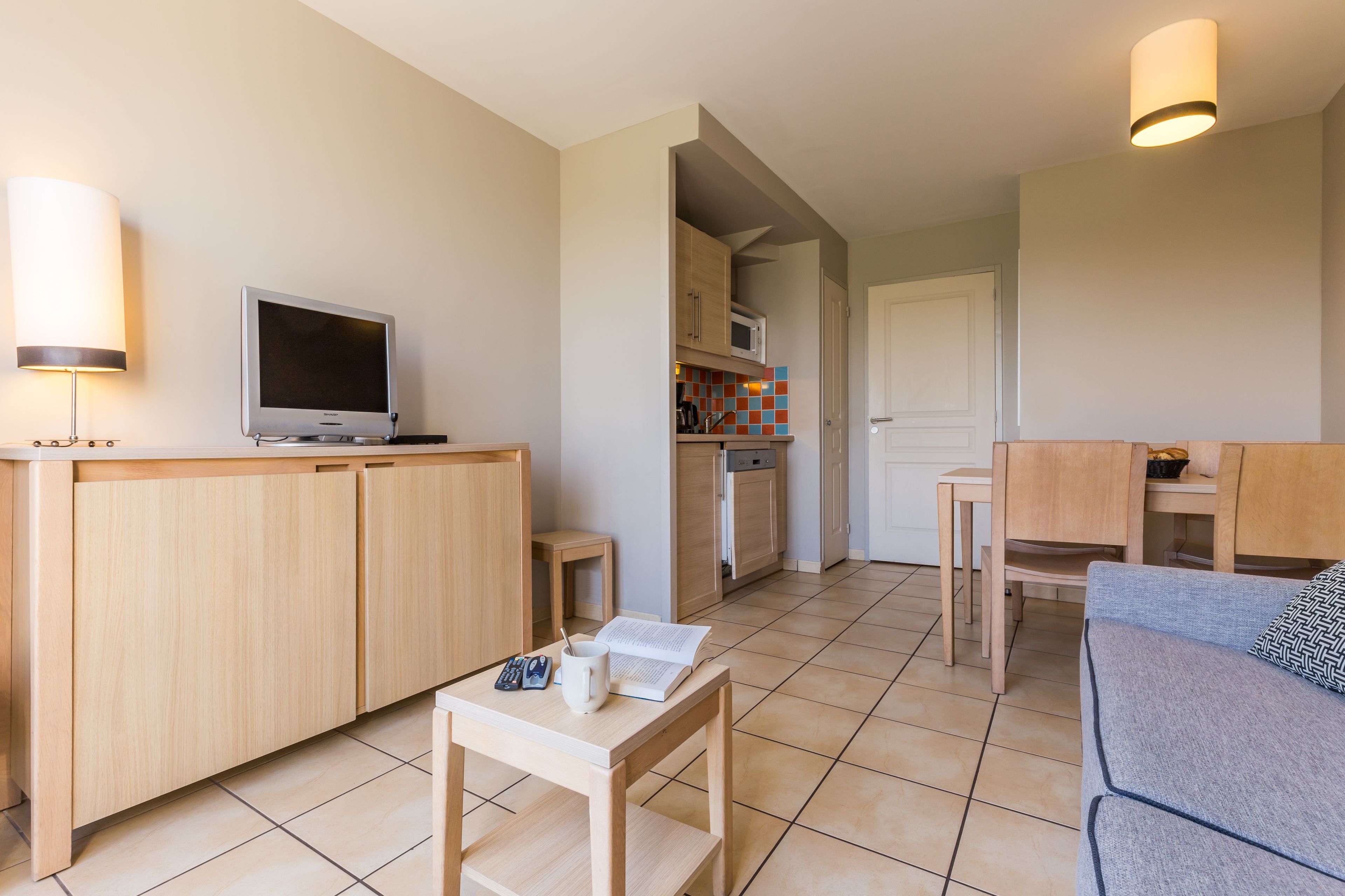 Una sala de estar moderna con televisor, una mesita con un libro y controles remotos, un sofá y una zona de cocina con alacenas y un microondas.
