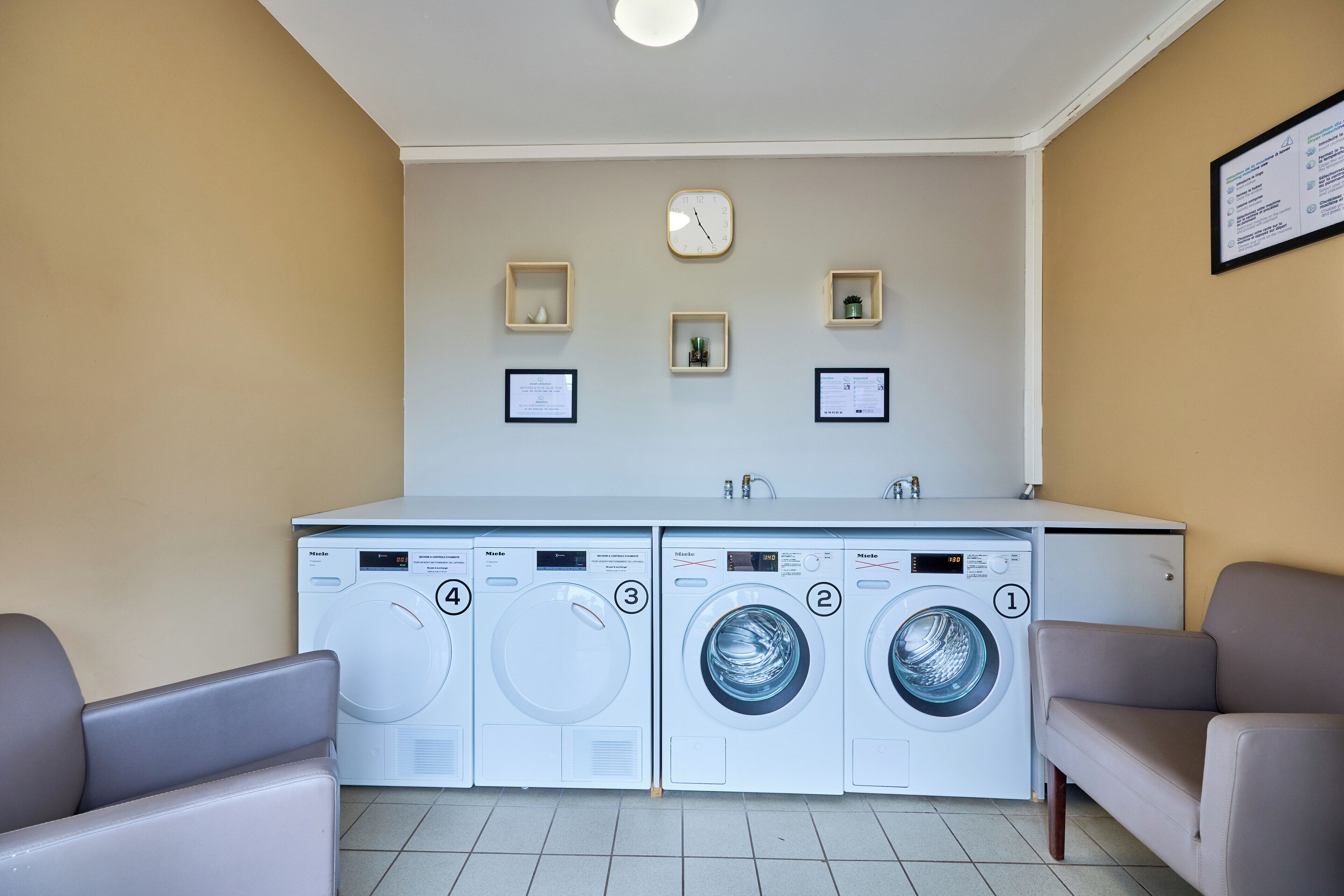 A laundry room with four washing machines, a clock, and framed certificates on the wall.