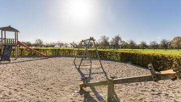 A playground with a swing set and a slide, surrounded by a sandy area and a hedge.