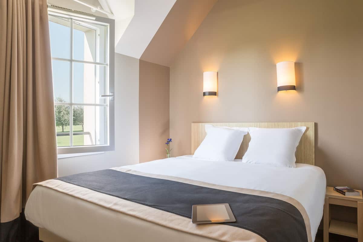 A hotel room with a large bed, two bedside lamps, a bedside table, and a window with curtains.