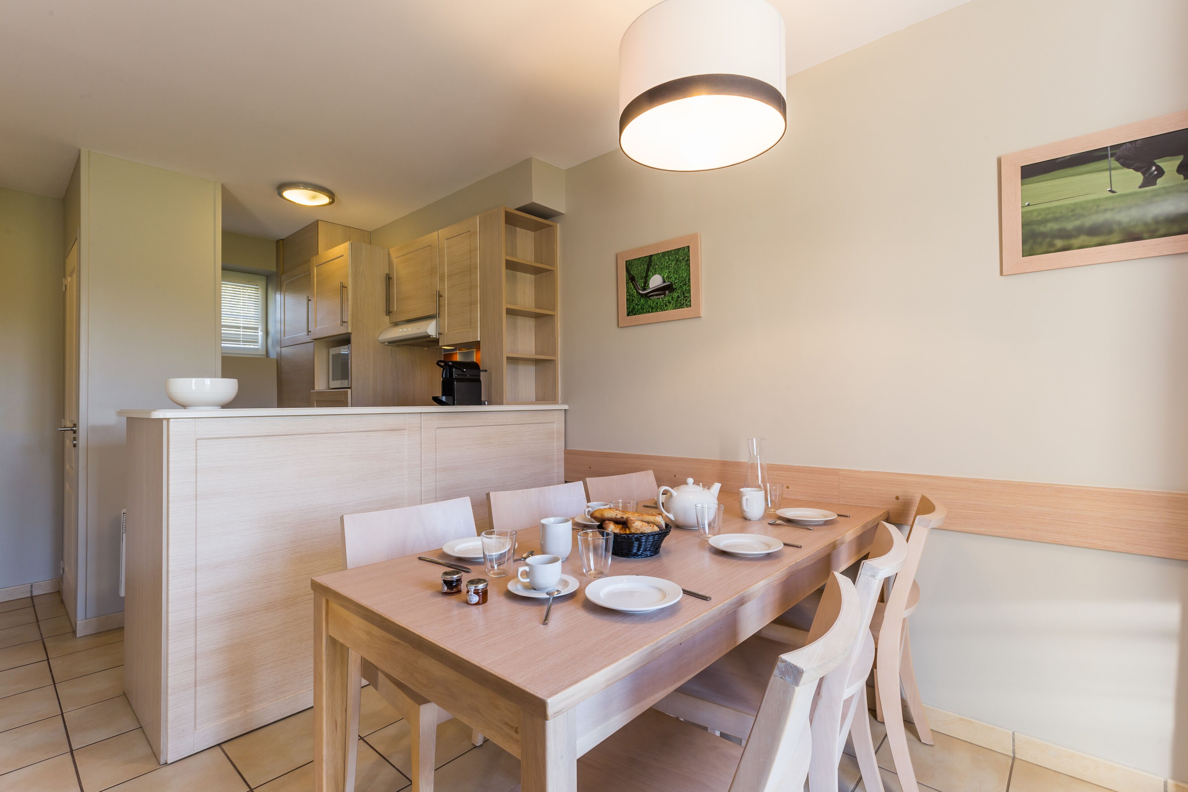 Un coin repas avec une table en bois dressée pour quatre personnes, une cuisine en arrière-plan et un tableau encadré au mur.