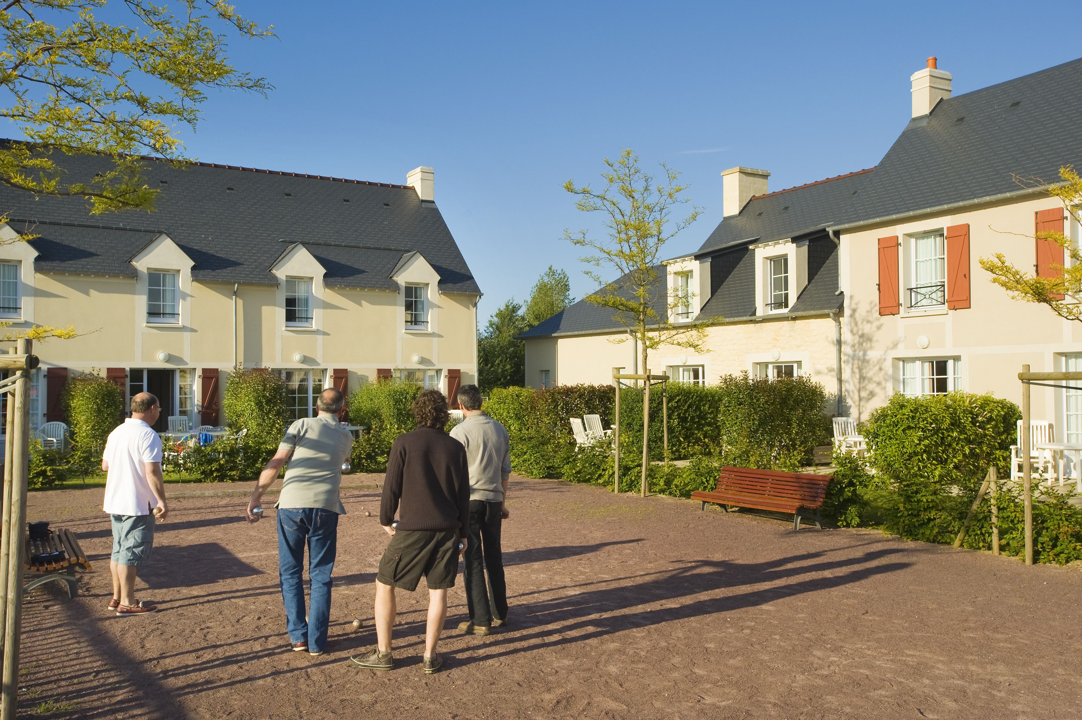 A courtyard with a dirt ground, a wooden bench, and a row of houses with red shutters and white trim.