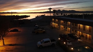 Un hôtel avec une piscine, plusieurs voitures garées devant, et une vue sur un coucher de soleil se reflétant sur une étendue d’eau.
