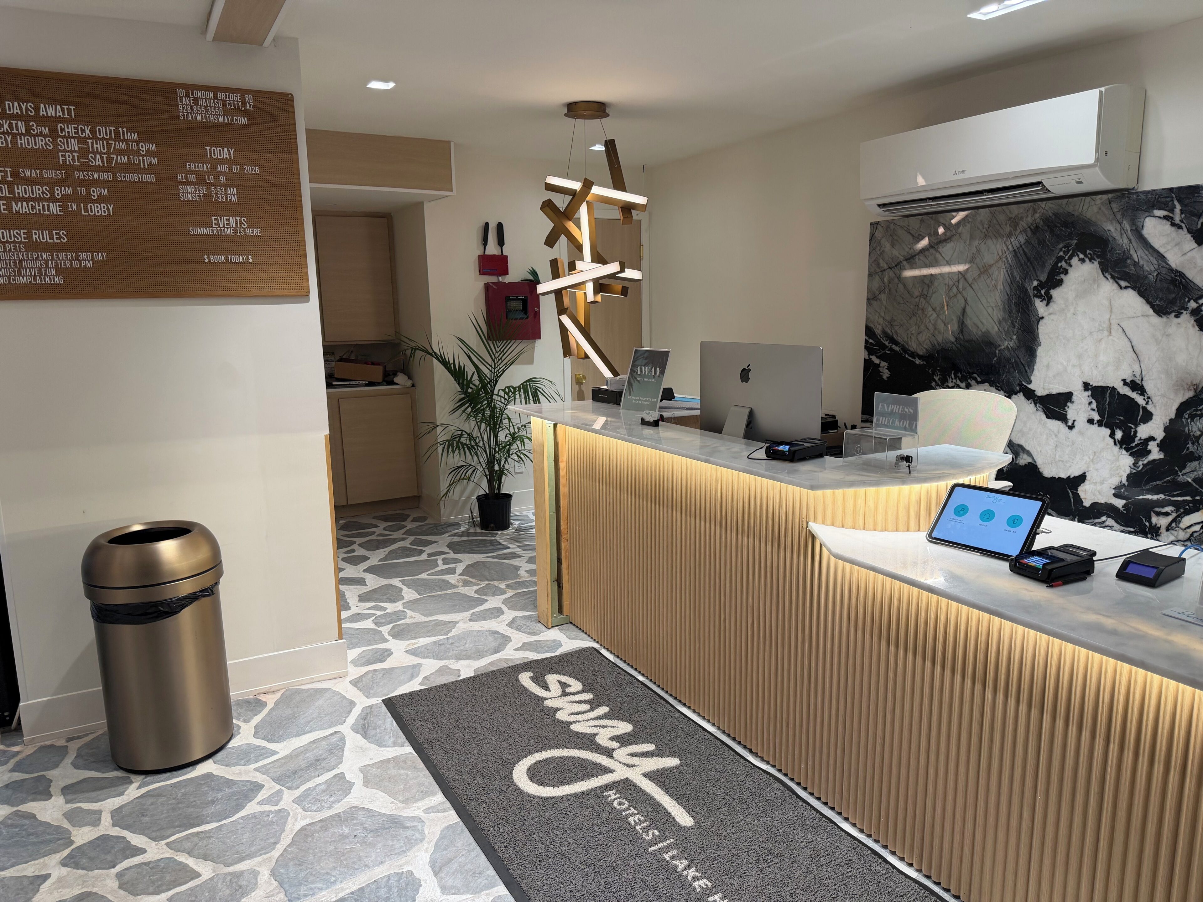 A hotel reception area with a stone-tiled floor, a reception desk with a computer and a phone, a trash can, and a wall-mounted sign with information.