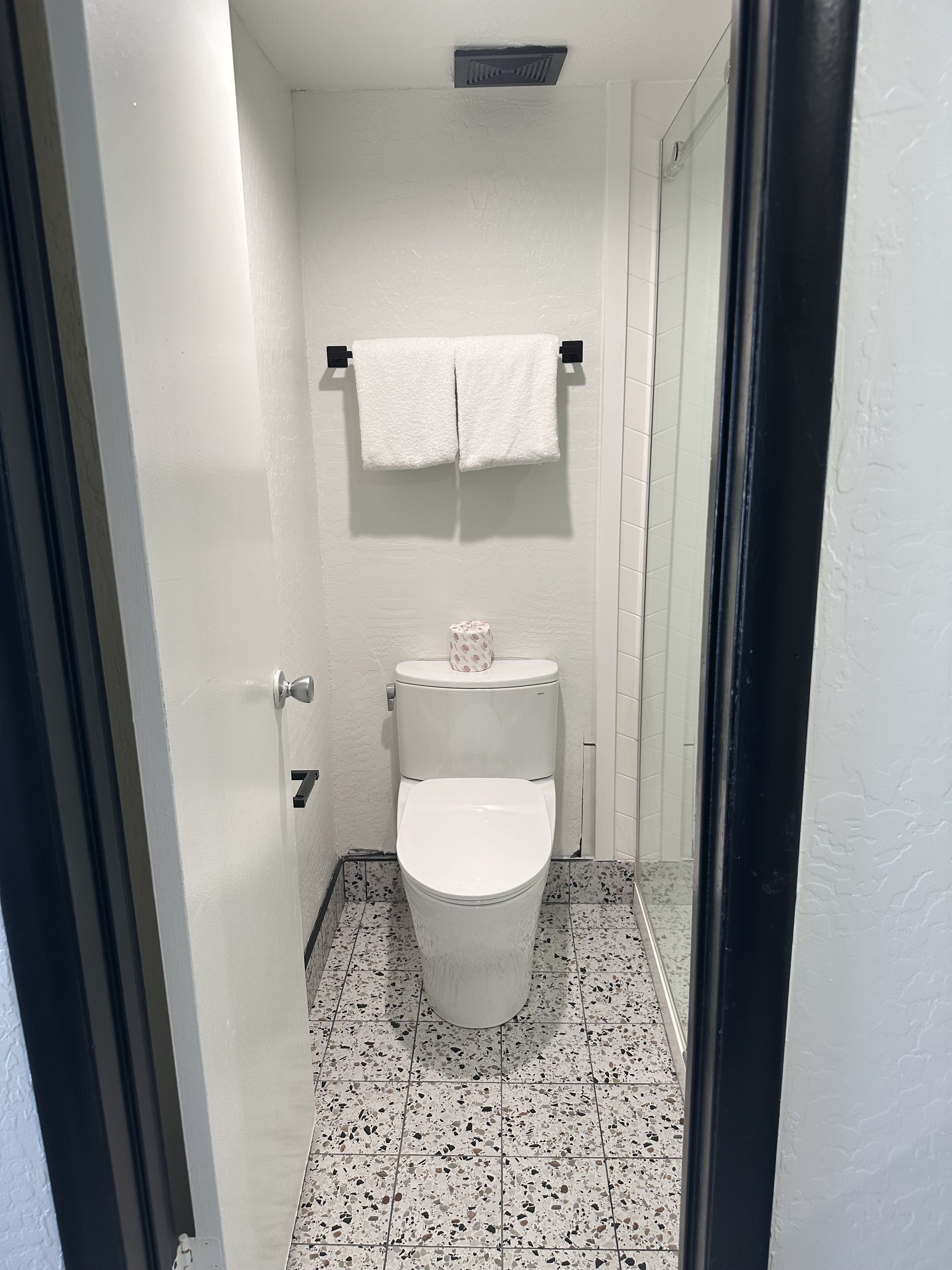 Casa de banho com sanita branca, um suporte de toalhas com duas toalhas e um rolo de papel higiénico.
