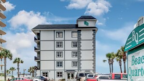 A multi-story hotel with balconies, a rooftop sign, and a parking area with cars.