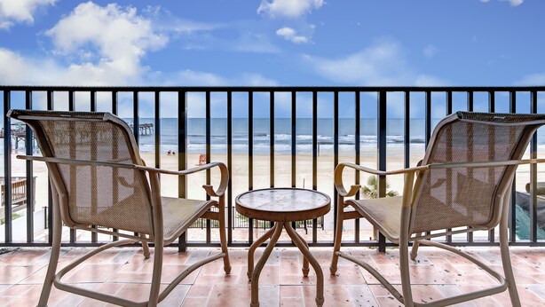 A balcony with two wicker chairs and a small round table, overlooking a beach with a clear sky and ocean.