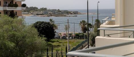 A coastal view from a balcony overlooking a road with parked cars, a streetlight, and a distant shoreline with buildings and a boat.