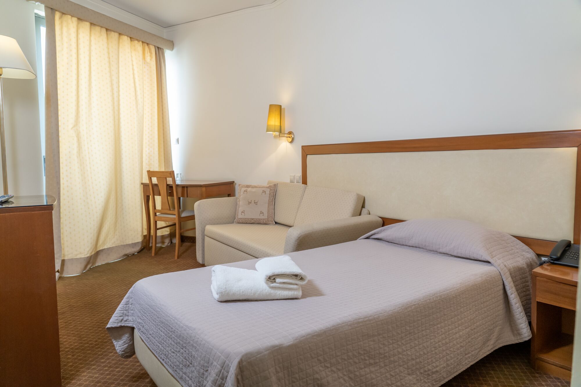 A hotel room with a bed, a desk, a chair, and a lamp.