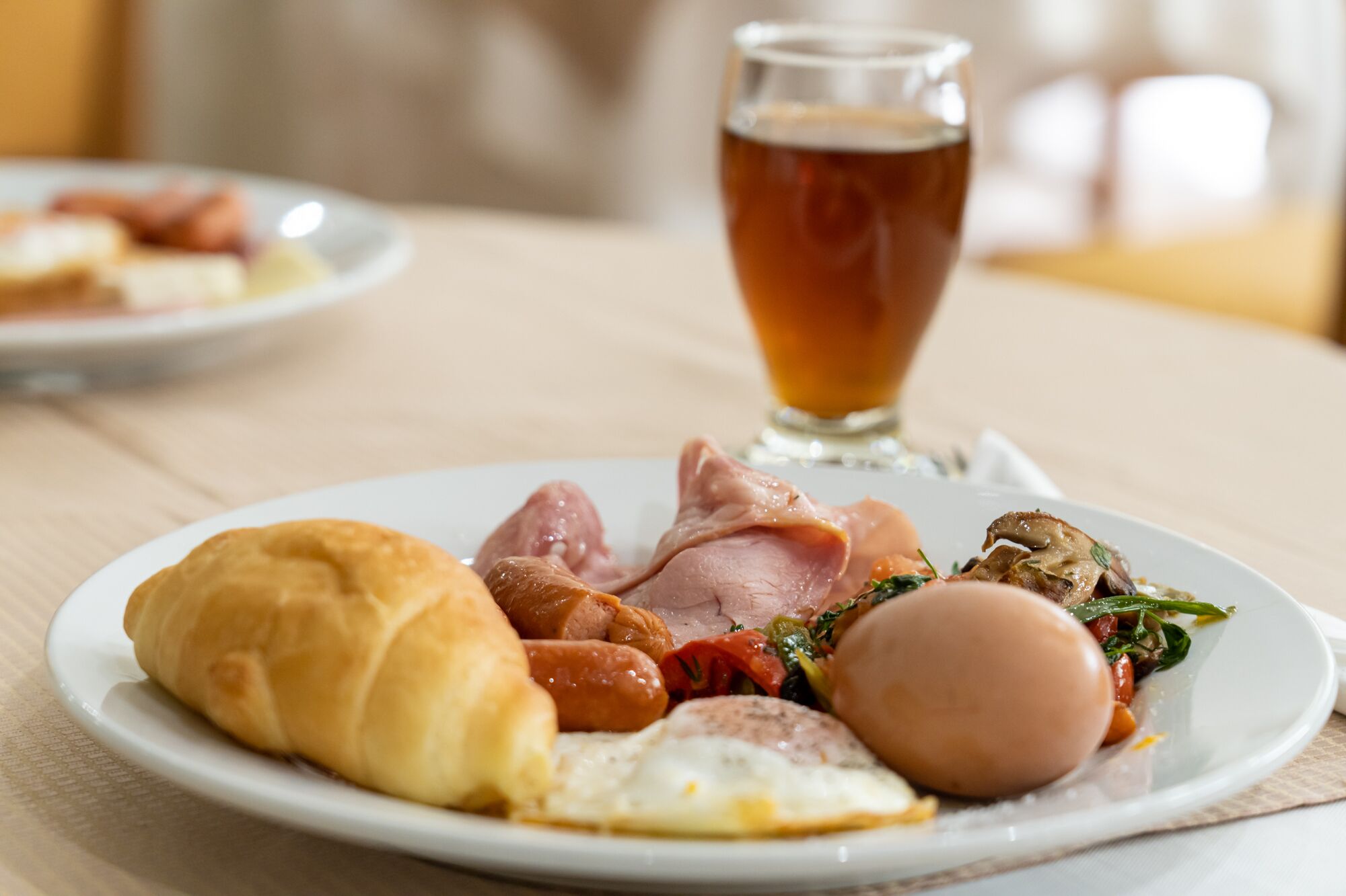 A plate with breakfast items including eggs, ham, sausages, and a croissant, with a glass of tea in the background.