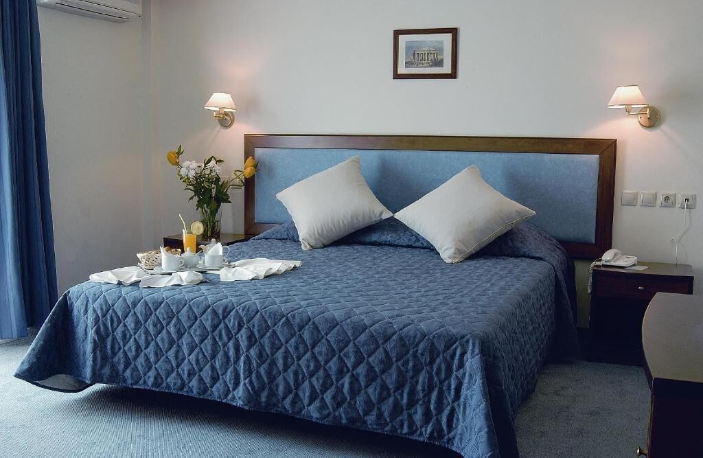 Una cama bien tendida con un edredón azul, dos almohadas, una mesita de noche con una lámpara y una pequeña mesa con cosas para el desayuno.