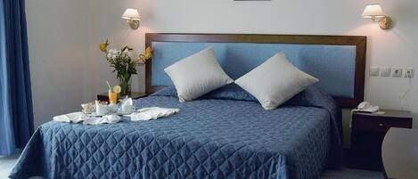 A neatly made bed with a blue quilt, two pillows, a bedside table with a lamp, and a small table with breakfast items.