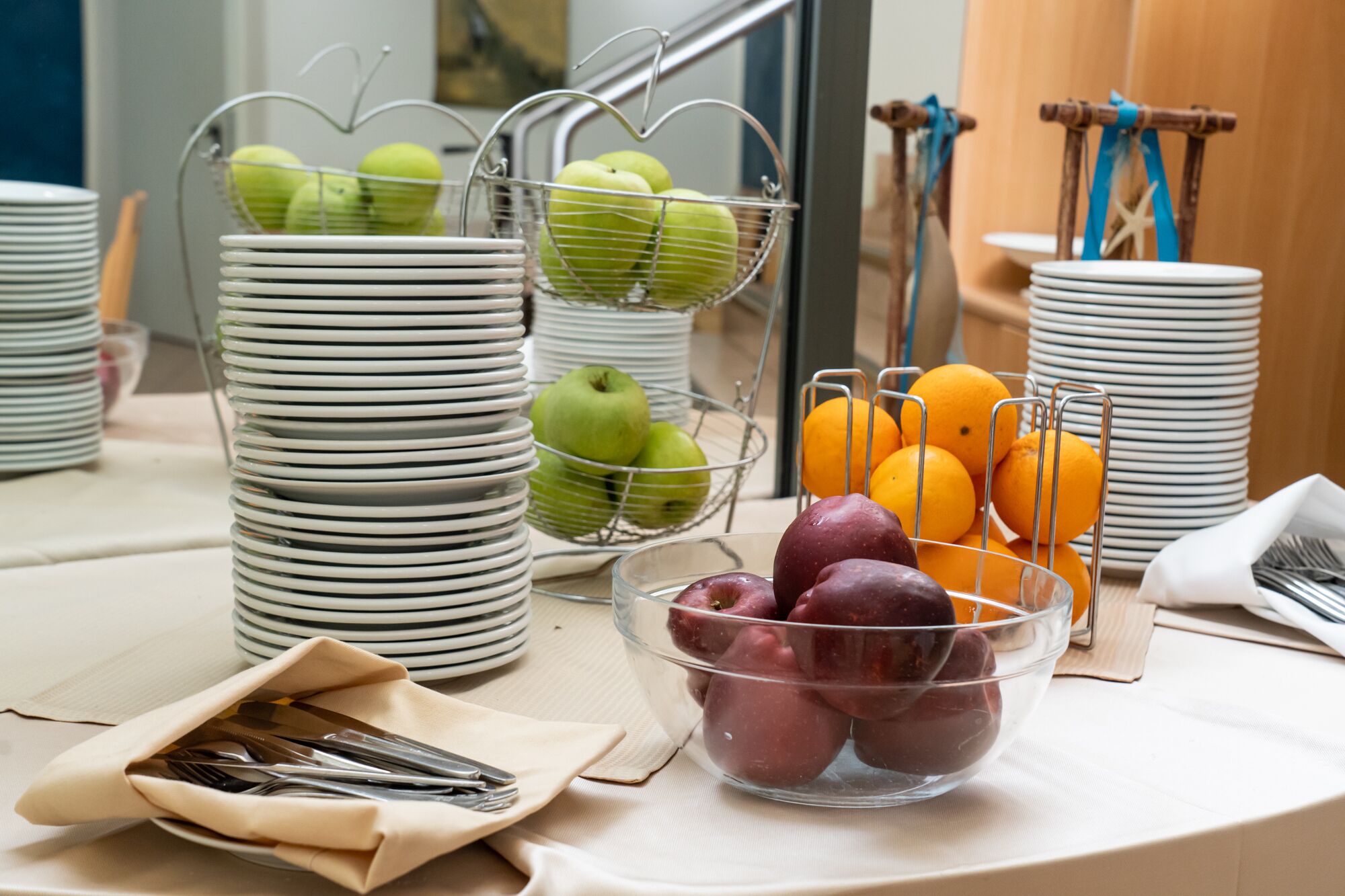 A breakfast buffet with stacks of white plates, a wire fruit basket with green apples, a bowl of red plums, and a wire fruit basket with oranges.